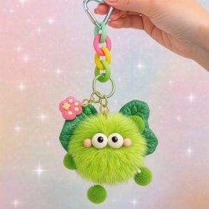 A hand holds a cute green fluffy keychain from the Charmies Keychain Collection by Pasha Love. The plush character has round eyes, pink cheeks, green wings, and colorful chain links in pink, yellow, and green with a blue heart-shaped clip. The background is soft pastel with sparkles, creating a dreamy and glam aesthetic.